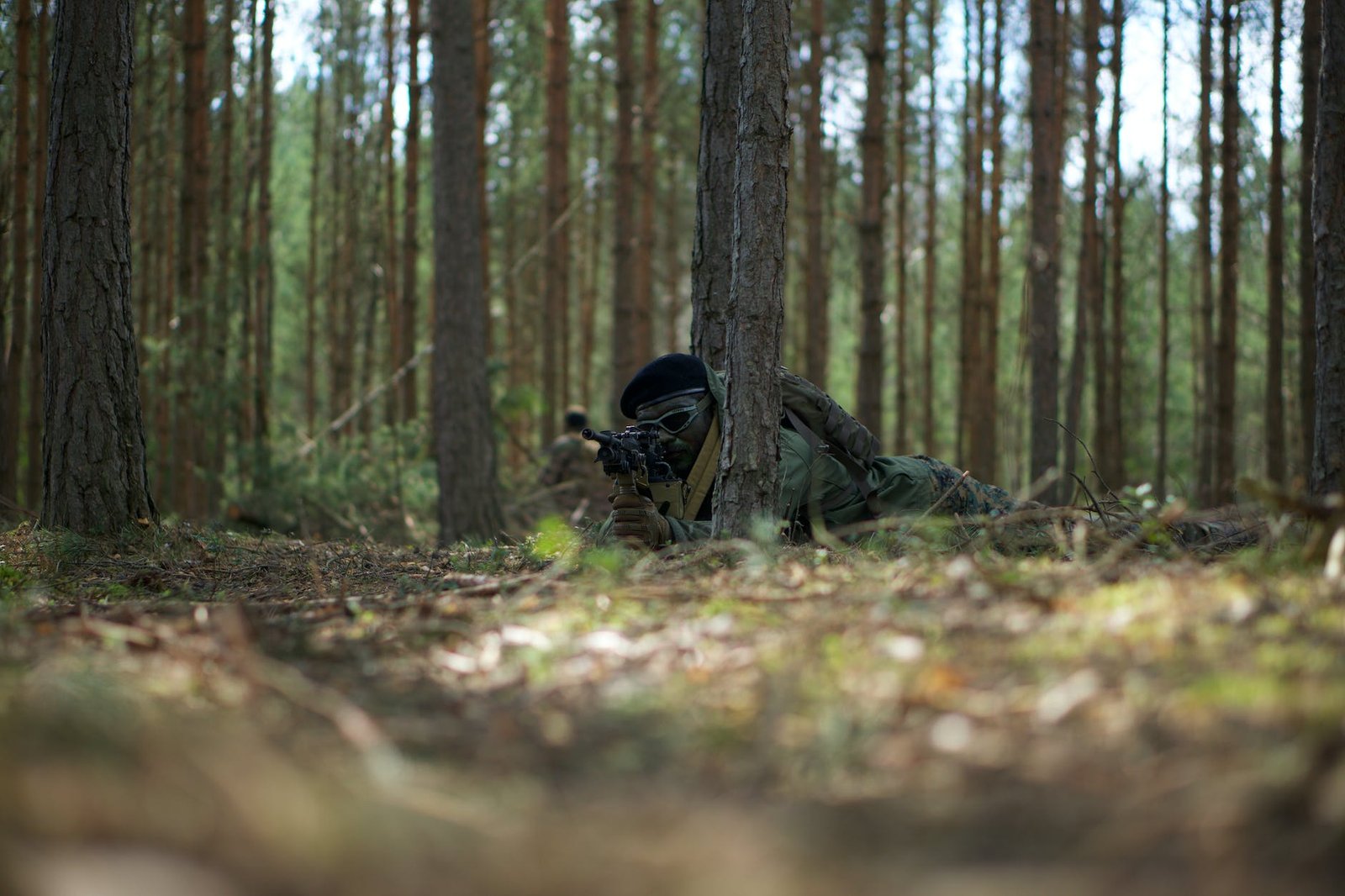man in military uniform and a backpack lying on ground surrounded by trees