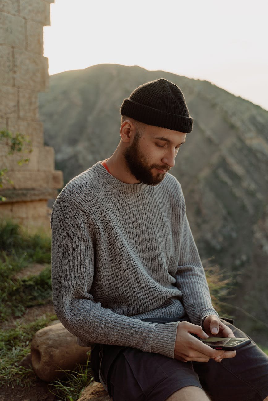man in gray sweater and black bonnet using cellphone with view of mountain on background
